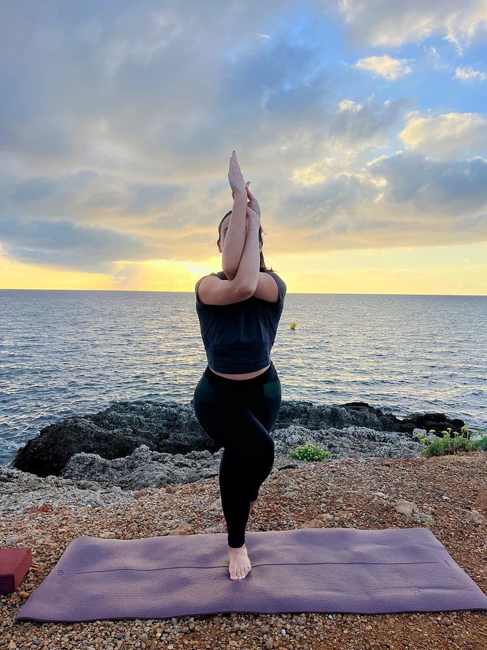 Jess en équilibre sur une jambe, les bras entrelacés devant le visage, au bord de la mer au coucher du soleil