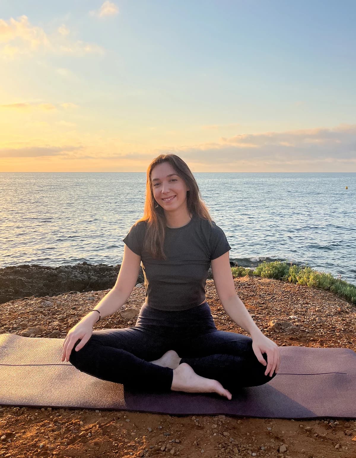 Jess smiling, sitting with her legs crossed on a yoga mat by the sea at sunset